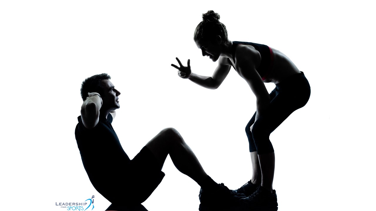 Ein Mann macht eine Sportübung und eine Frau zeigt mit ihren Fingern an, wie viele Ausführungen er noch machen muss.